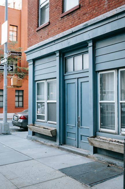 A front exterior view of a blue-painted building with large white-framed windows and a central door, situated on a narrow street in Greenwich. The building has horizontal wooden cladding and decorativ