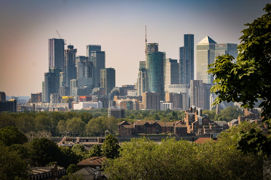 A view of a modern city skyline with numerous tall skyscrapers and high-rise office buildings under a partly cloudy sky, seen from a park with lush green trees and well-maintained grass in the foregro