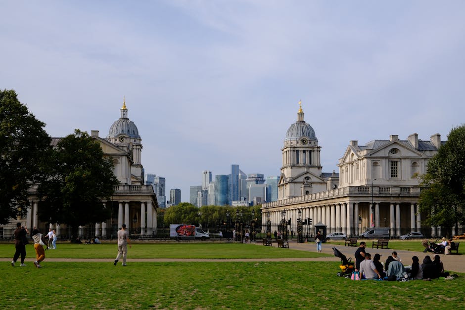 An expansive view of the Royal Palace of Greenwich, with its white classical façade featuring columns and decorative architectural details, is visible in the foreground. Surrounding the palace is a w