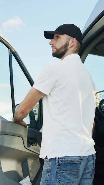A young man with a beard and wearing a white polo shirt and black cap is standing outside a box truck on a driveway or pavement. He is holding a cardboard box and smiling at the camera. The truck’s